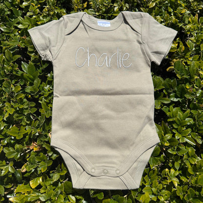 Sage green baby onesie with 'Charlie' printed on it, lying on green foliage.