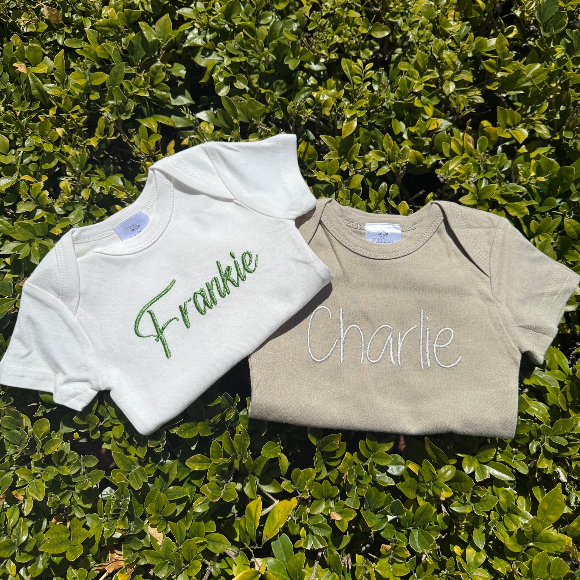 Two baby onesies with embroidered names 'Frankie' and 'Charlie' on a green leafy background.
