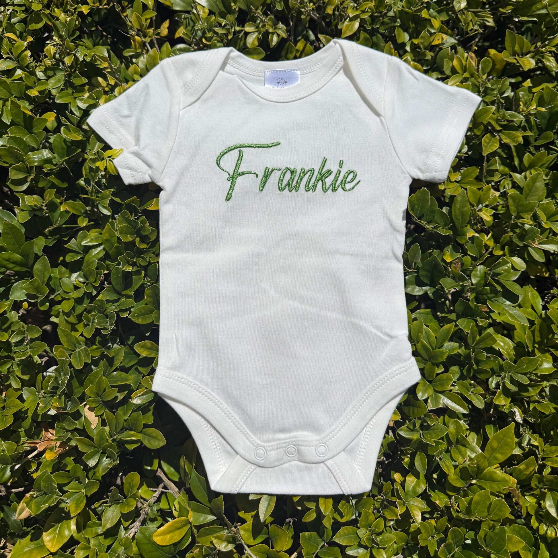 White baby onesie with 'Frankie' printed on it, lying on green foliage.